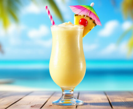 Virgin Colada servita in un bicchiere hurricane, decorata con fetta di ananas, ciliegina rossa e ombrellino colorato, su tavolo di legno davanti al mare tropicale.