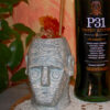 Cocktail Nuragic Echo servito in un bicchiere artistico ispirato ai Giganti di Mont’e Prama, accanto a una bottiglia di P31 Green Bitter, su tovaglietta di pizzo, con sfondo caldo e pianta decorativa. Questa ricetta cocktail ha vinto la prima edizione della P31 Cocktail Kombat