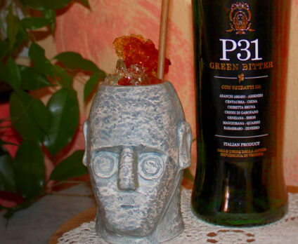 Cocktail Nuragic Echo servito in un bicchiere artistico ispirato ai Giganti di Mont’e Prama, accanto a una bottiglia di P31 Green Bitter, su tovaglietta di pizzo, con sfondo caldo e pianta decorativa. Questa ricetta cocktail ha vinto la prima edizione della P31 Cocktail Kombat