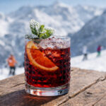 Cocktail Milano Cortina servito in tumbler basso con menta spolverata di zucchero a velo, fetta di arancia e sfondo innevato di montagna