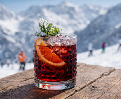 Cocktail Milano Cortina servito in tumbler basso con menta spolverata di zucchero a velo, fetta di arancia e sfondo innevato di montagna